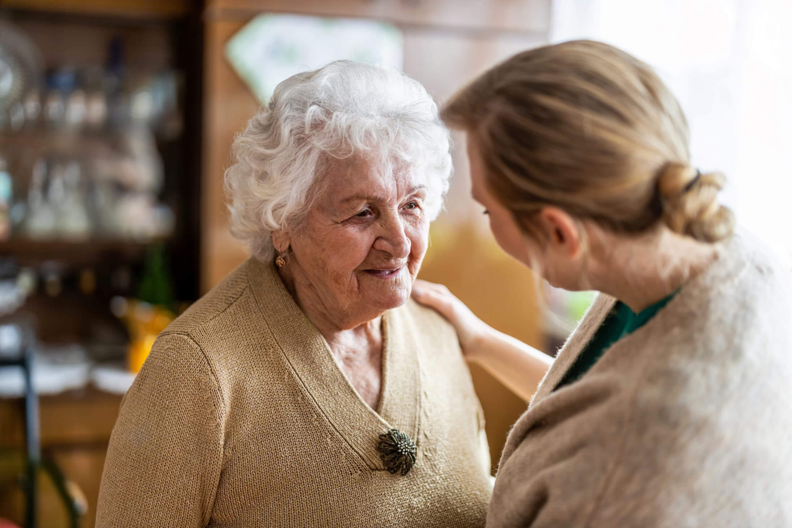 Caregiver facing an elderly woman with a warm, attentive expression, hand on her shoulder