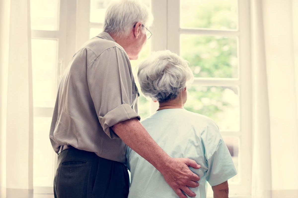 Elderly couple standing together at home, looking out a bright window